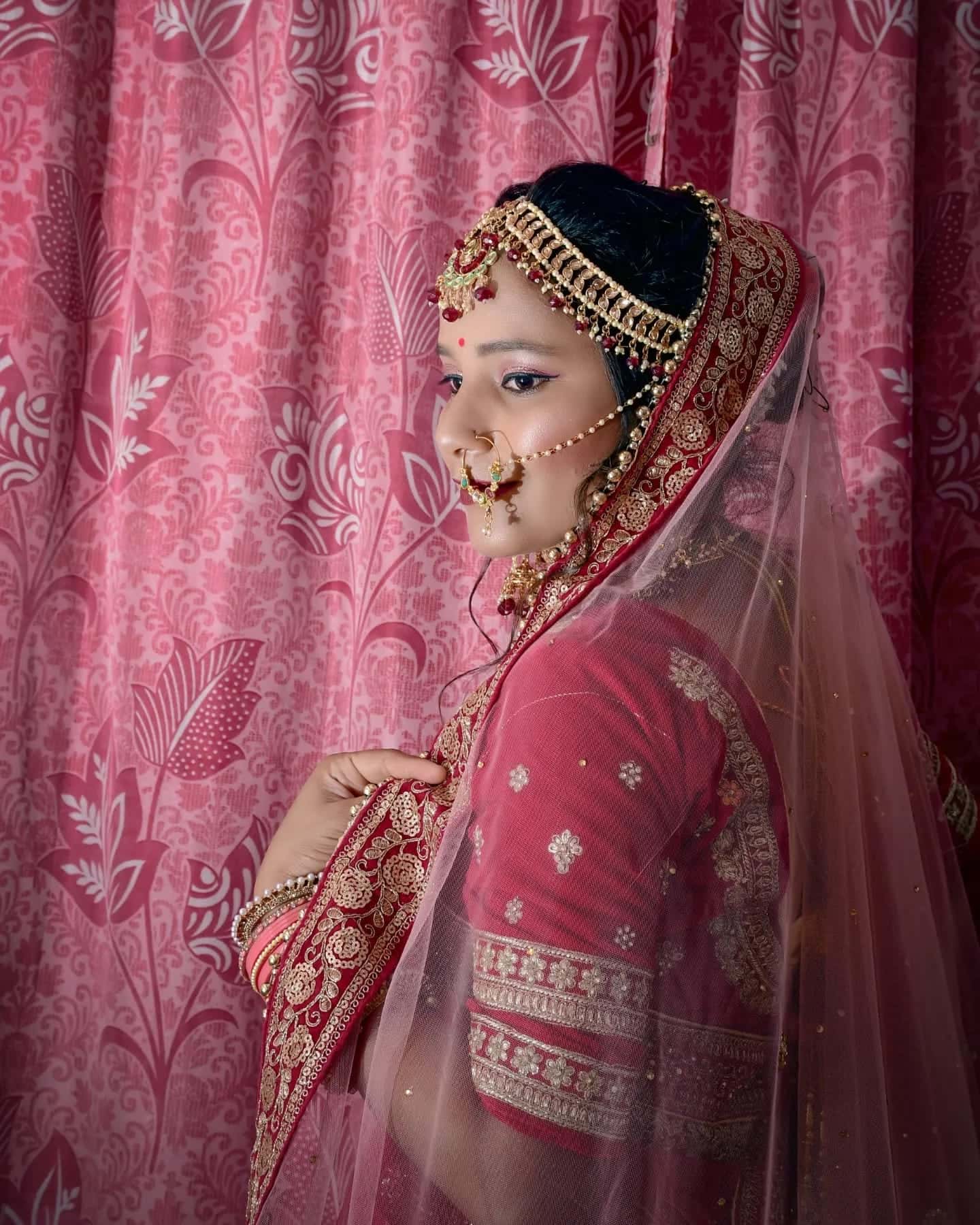 A bride in profile in a red lehenga with a sheer veil and gold matha patti