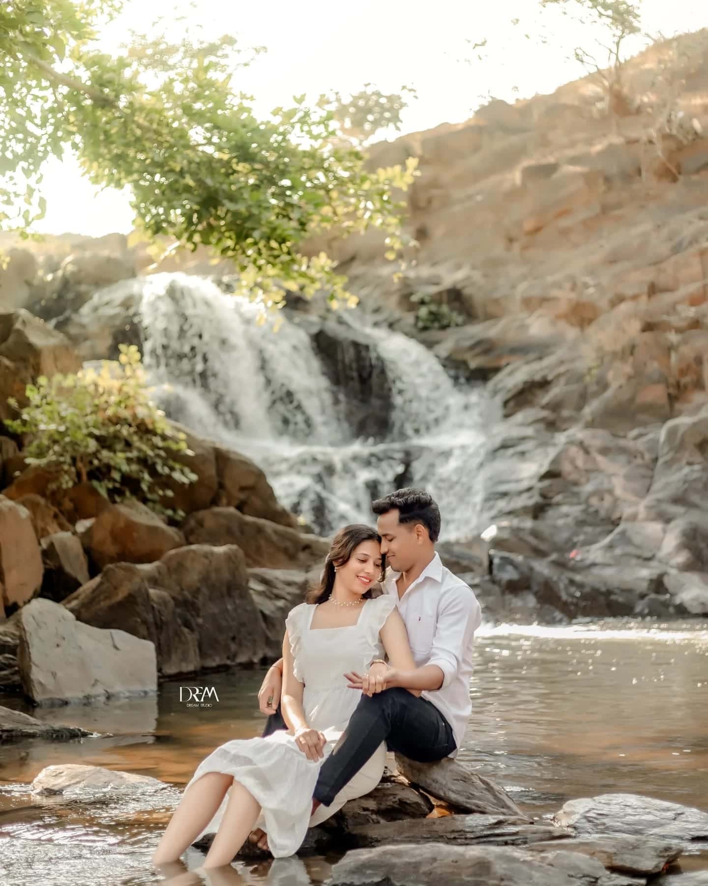 A couple in white sit on rocks in a stream with a waterfall behind them