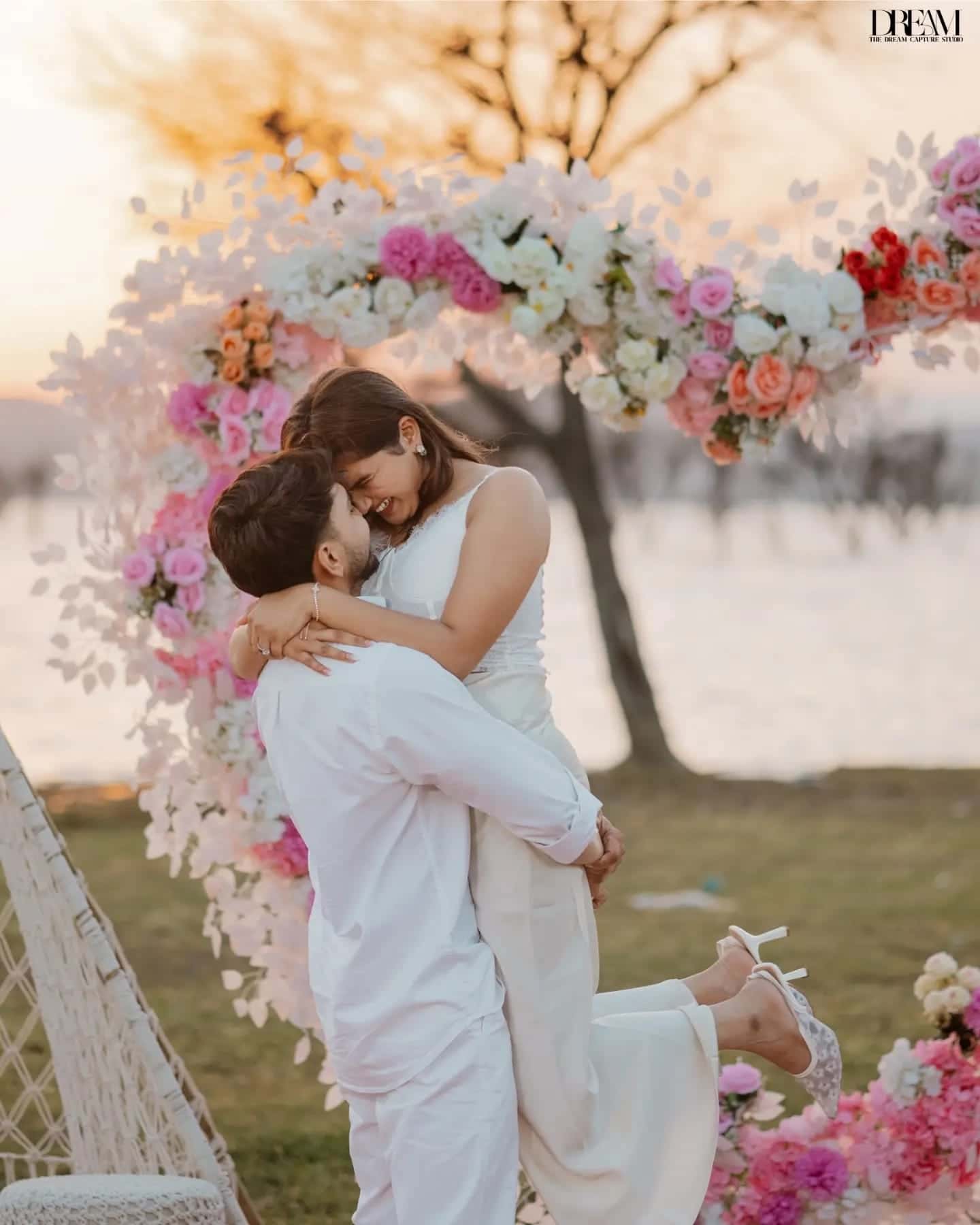 A man lifts a laughing woman in a white gown in front of a floral arch at golden hour
