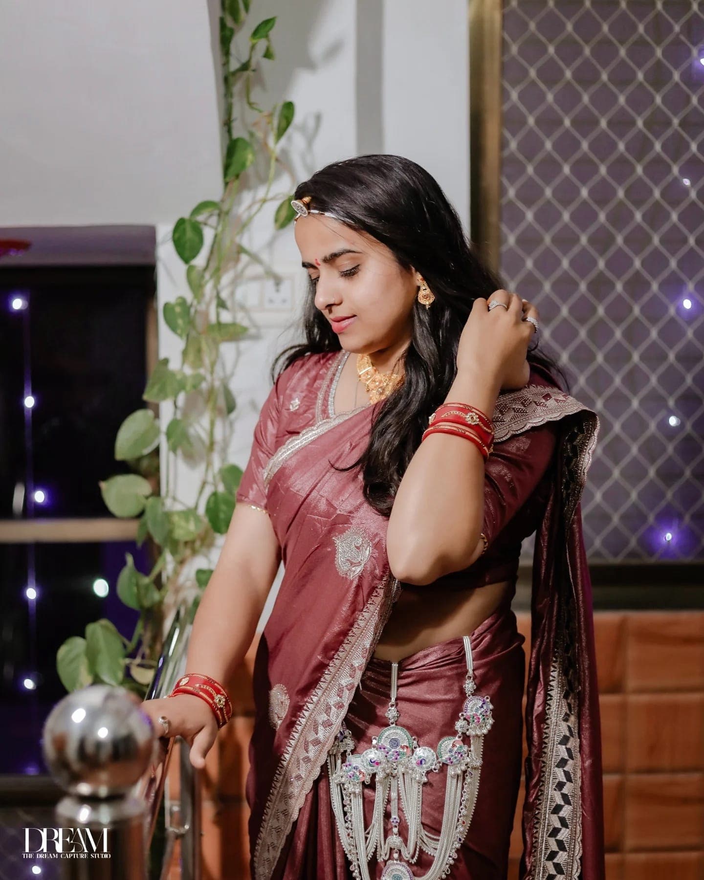 A bride in a maroon silk saree looks down, one hand in her hair