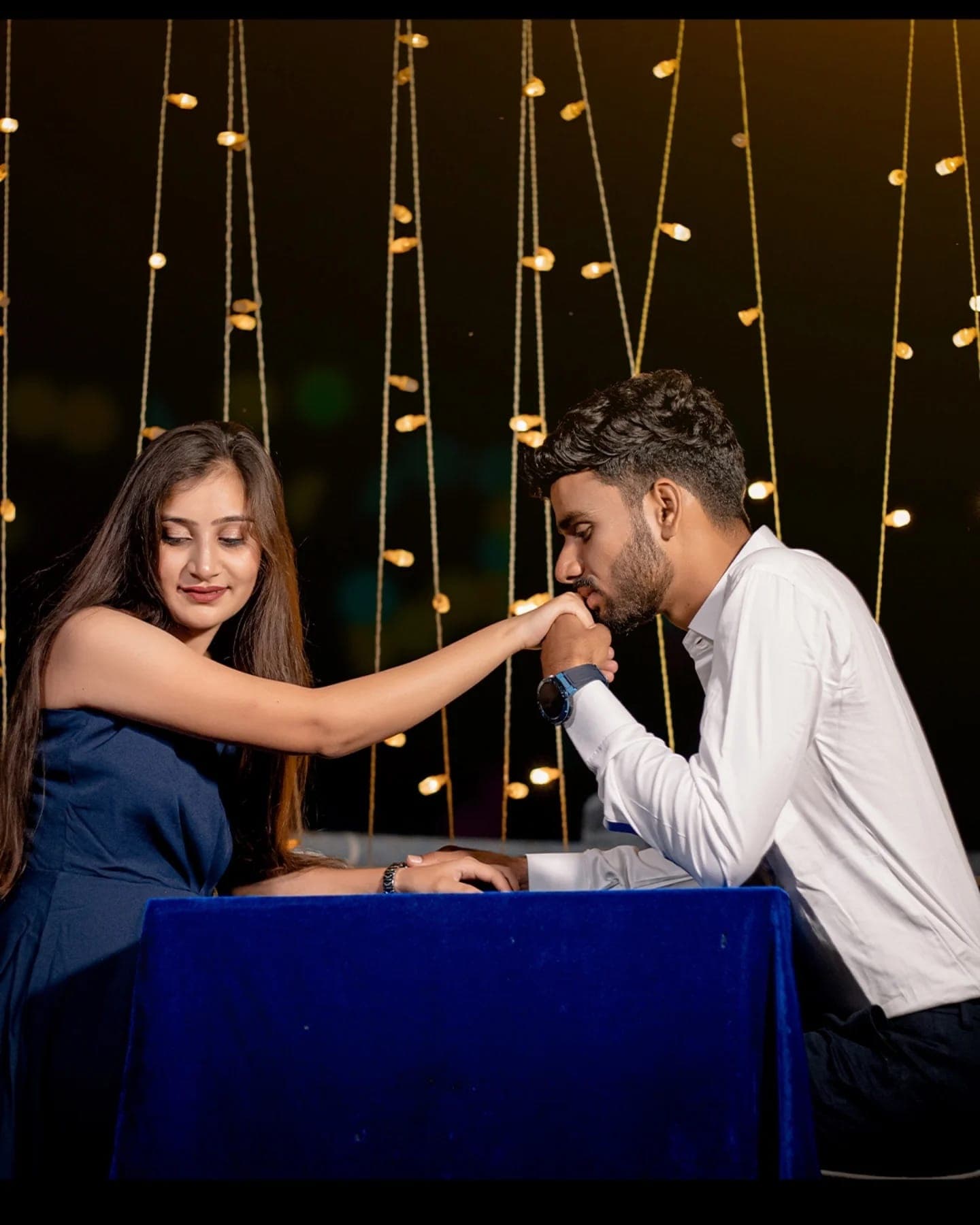 A man kisses a woman’s hand across a velvet table under hanging string lights