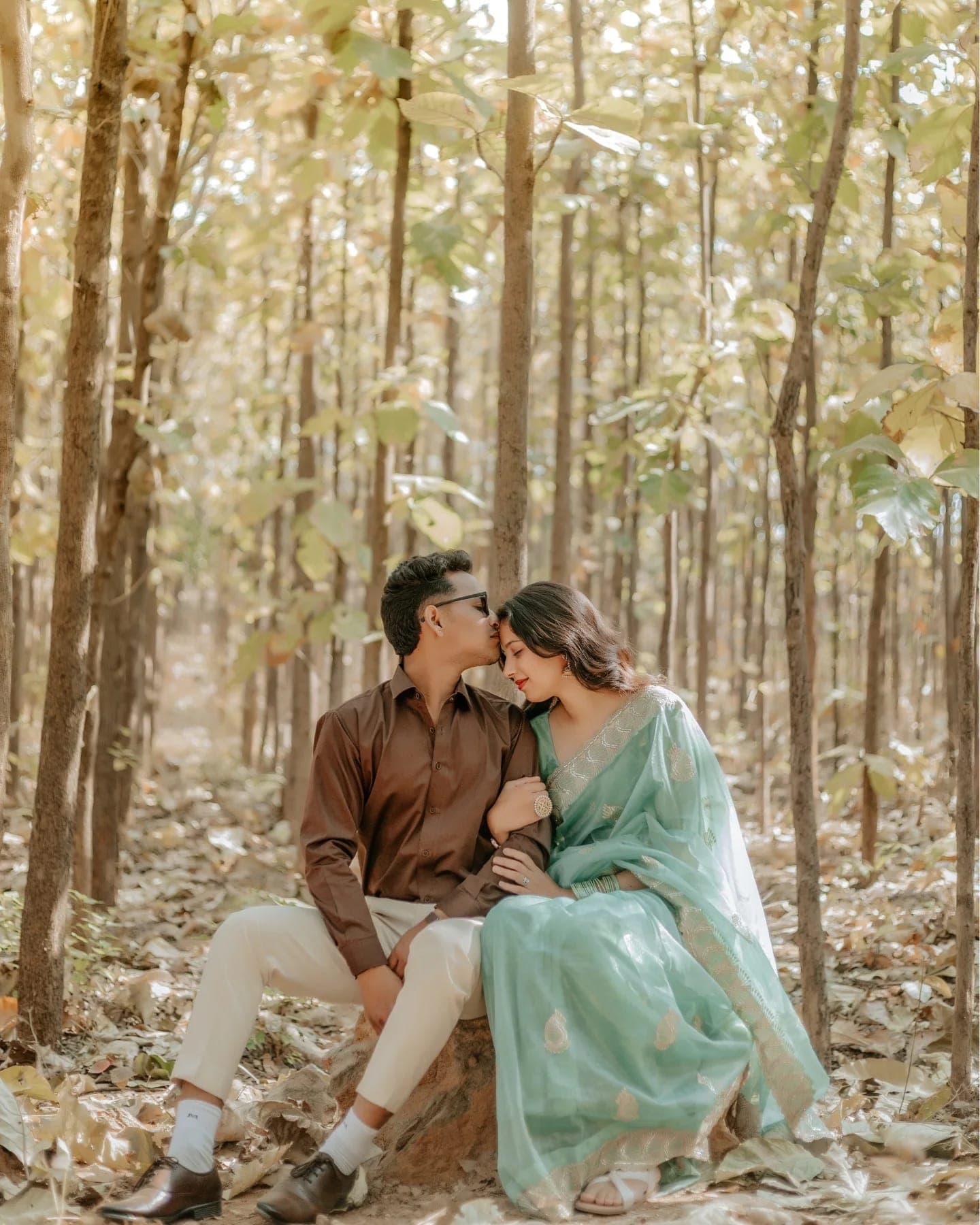 In a sunlit teak forest, a man kisses the forehead of a woman in a mint saree