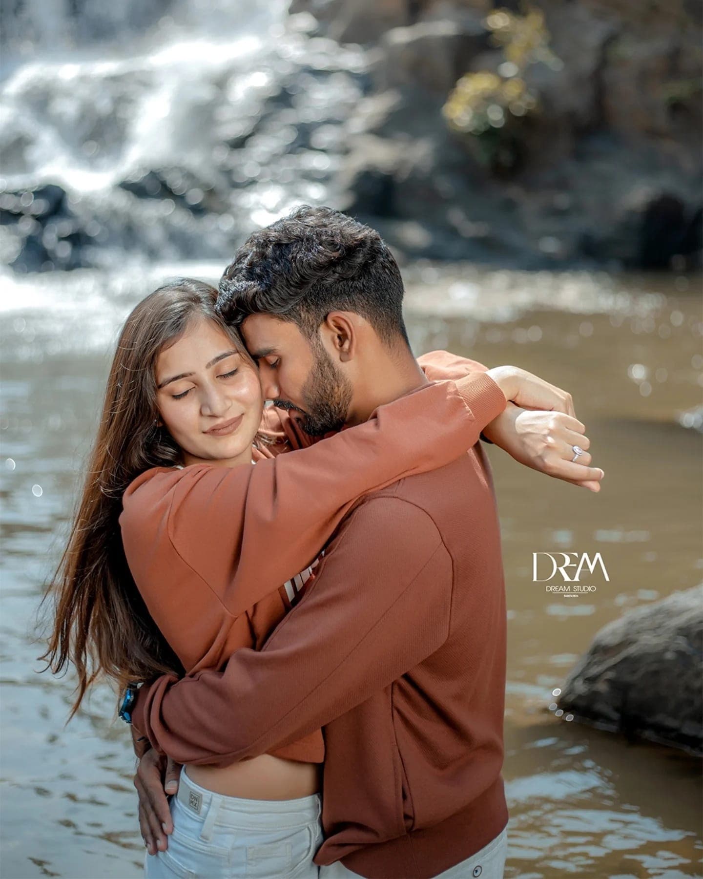 A couple in matching rust sweaters embrace with eyes closed, standing in a stream