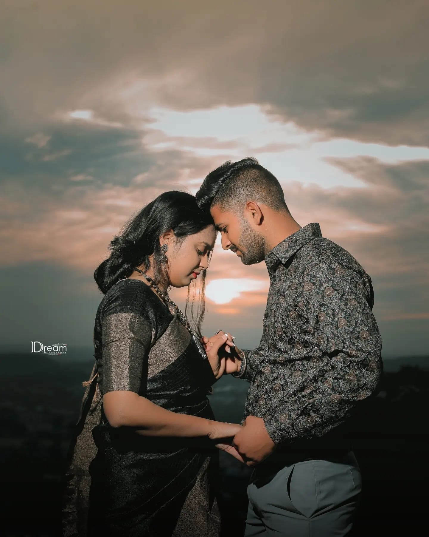 A couple hold hands forehead to forehead against a sunset sky on a hilltop