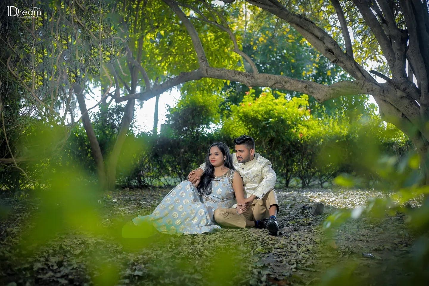 A couple sit together on the forest floor framed by green branches