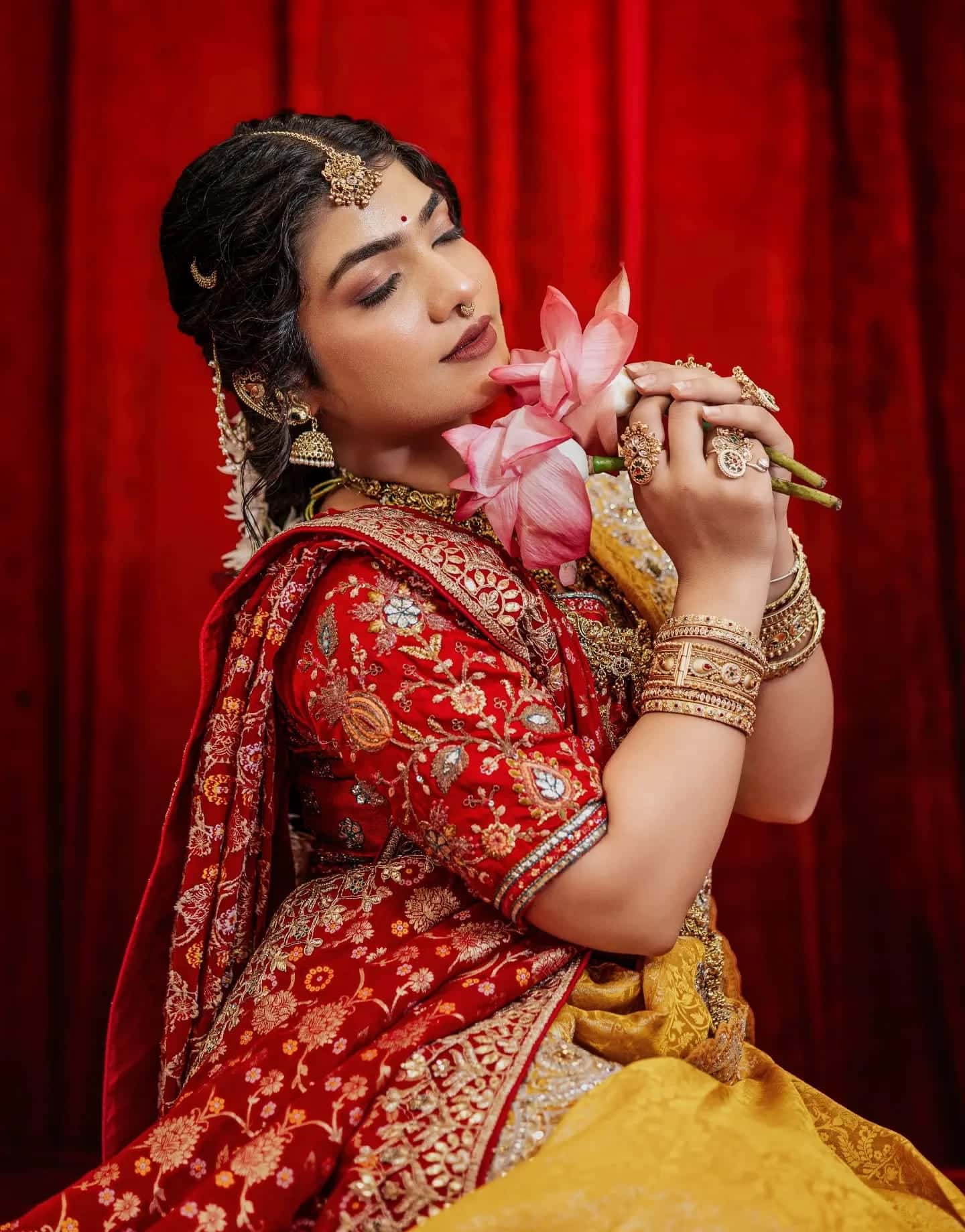 A model in red bridal wear holds pink lotus flowers to her face, eyes closed, against a red curtain