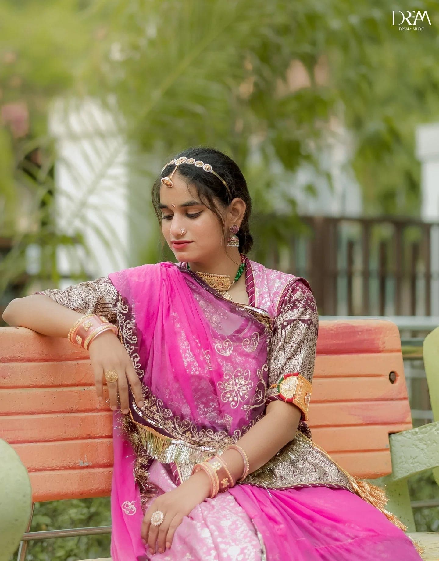 A young woman in a pink and gold Rajasthani poshak sits on a bench, looking down