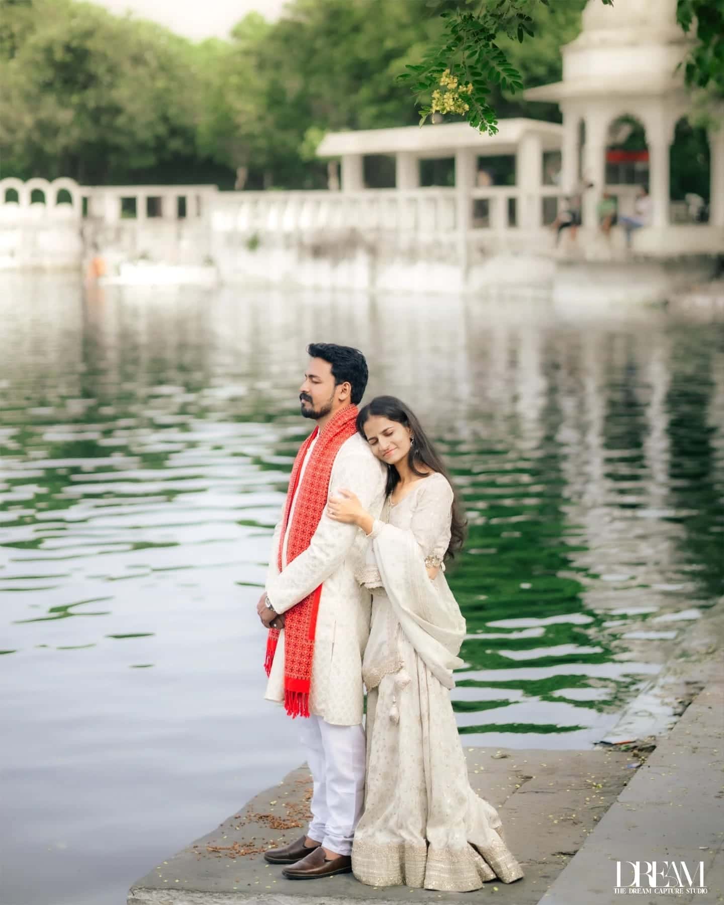 A couple in ivory outfits and a red stole stand on a white marble lakeside ghat, her head resting on his shoulder