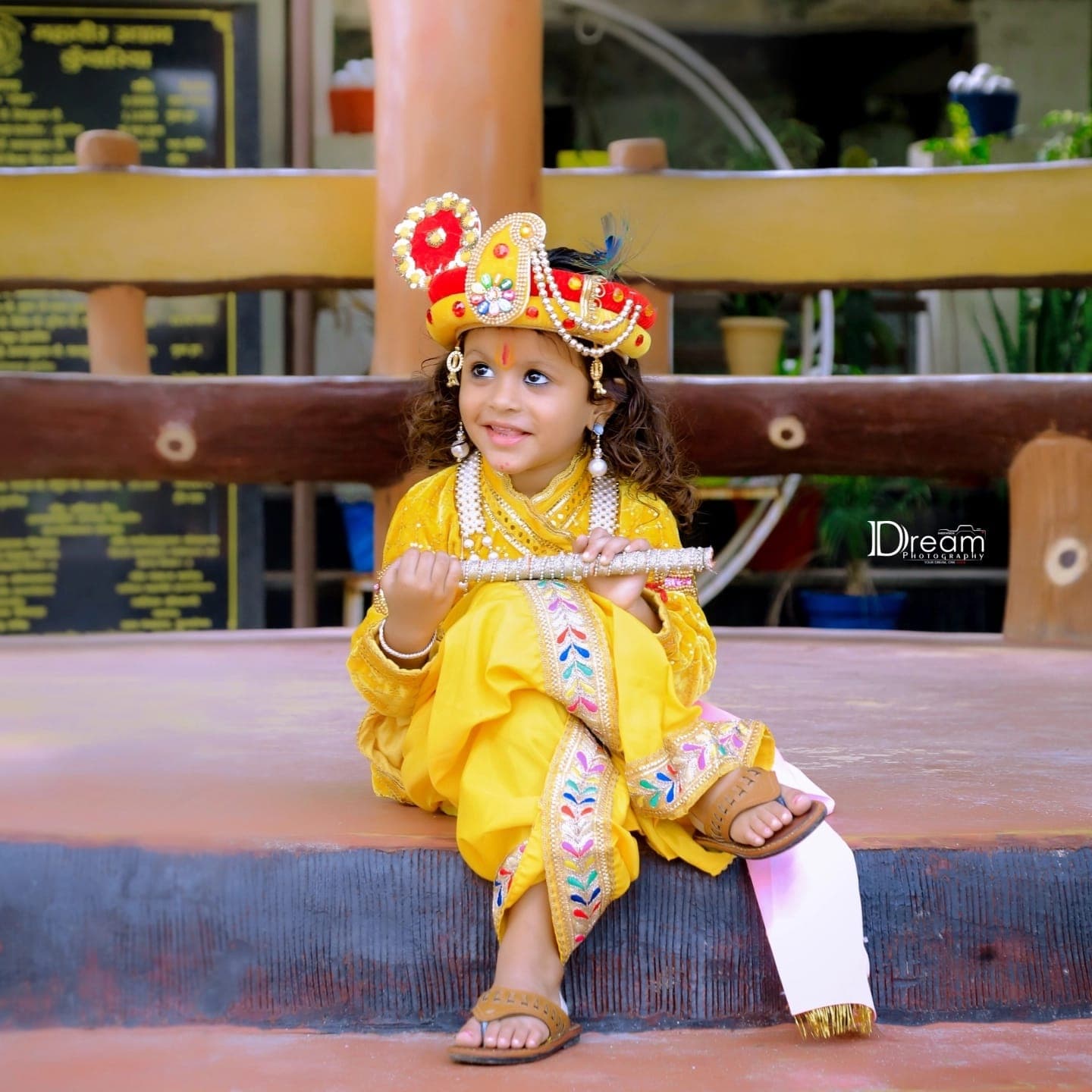 A smiling child in a Krishna costume and jewelled crown holds a silver flute