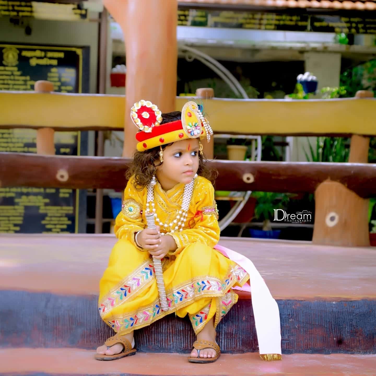 A small child dressed as Krishna in yellow sits on a step holding a flute