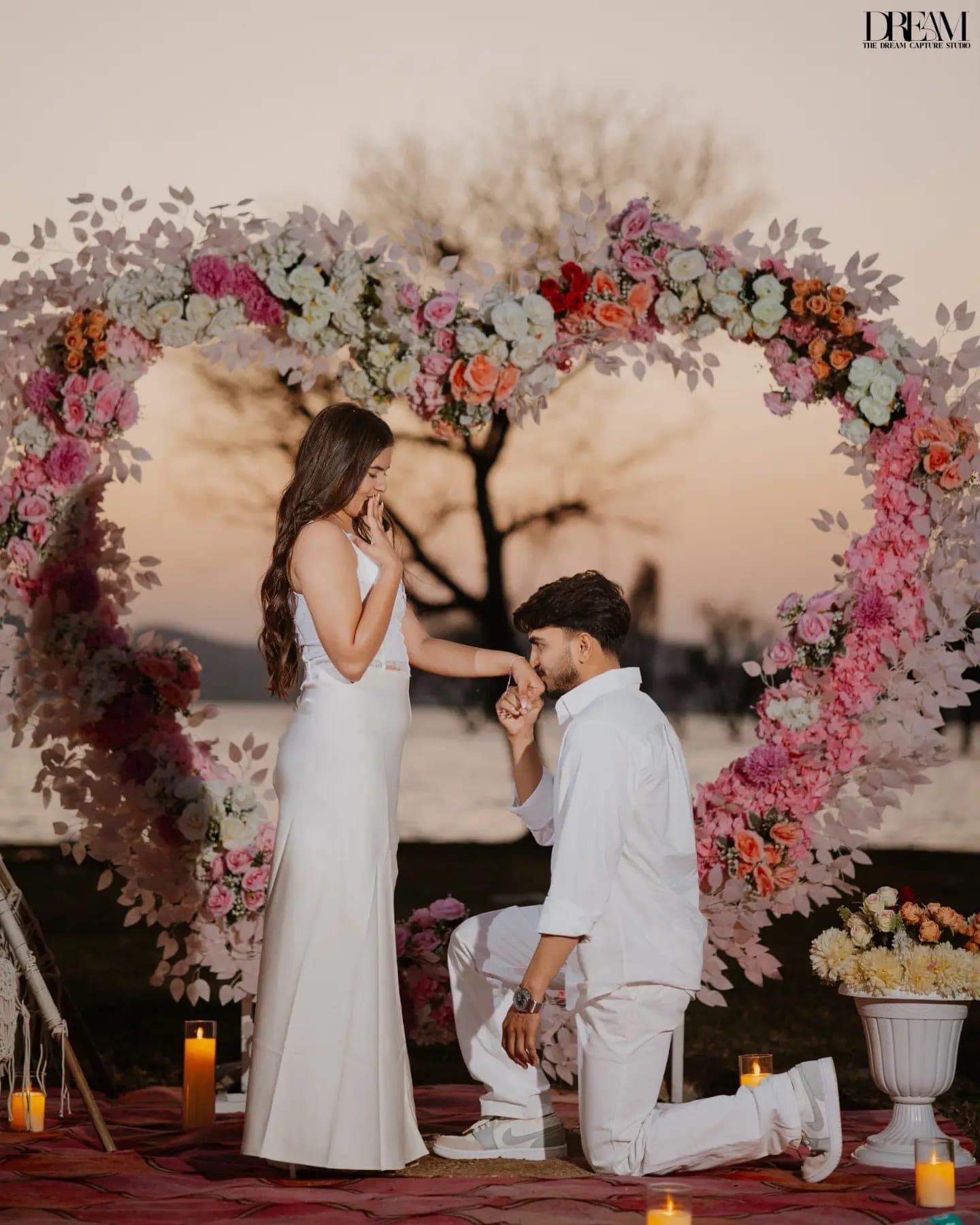 Inside a floral heart arch, a kneeling man kisses a woman’s hand