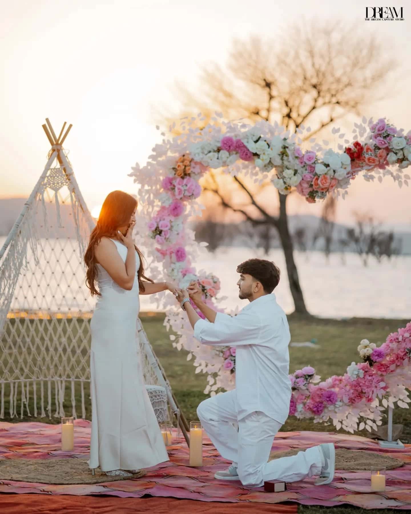 A man kneels to propose beside a pink floral heart arch by a lake