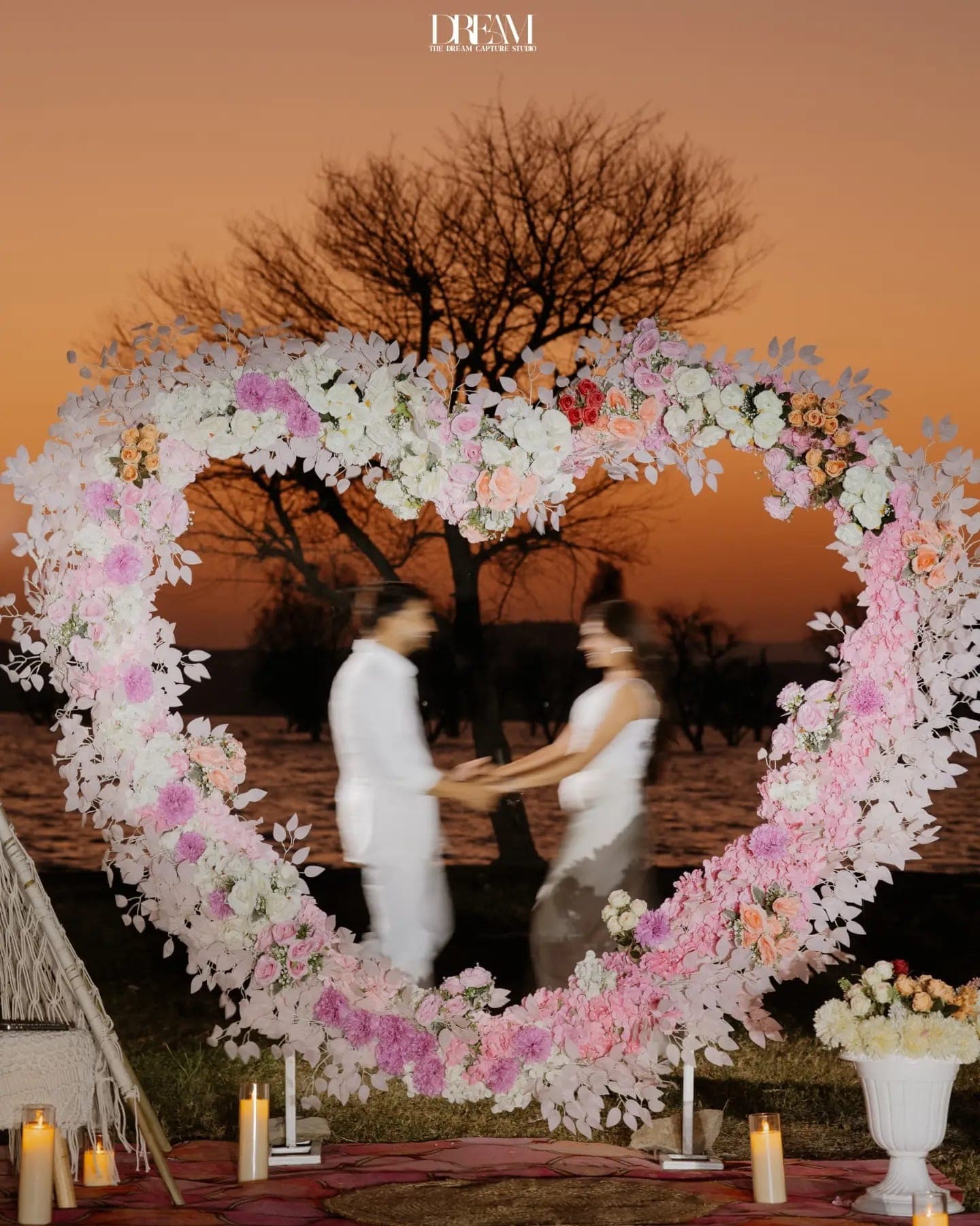 A couple hold hands inside a floral heart arch against an orange sky
